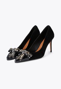 Talons hauts en suede noir avec un bout pointu et un accent en vernis brillant. Un nœud décoratif présente des embellissements métalliques mixtes. Talon aiguille haut.