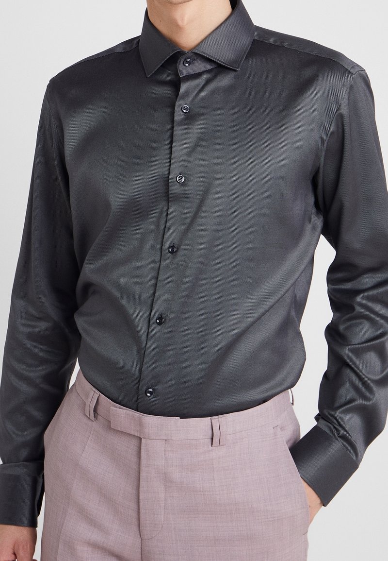 Camicia grigia scura a maniche lunghe realizzata in tessuto liscio con una leggera lucentezza. Presenta un colletto classico e bottoni scuri. Abbinata a pantaloni chiari.