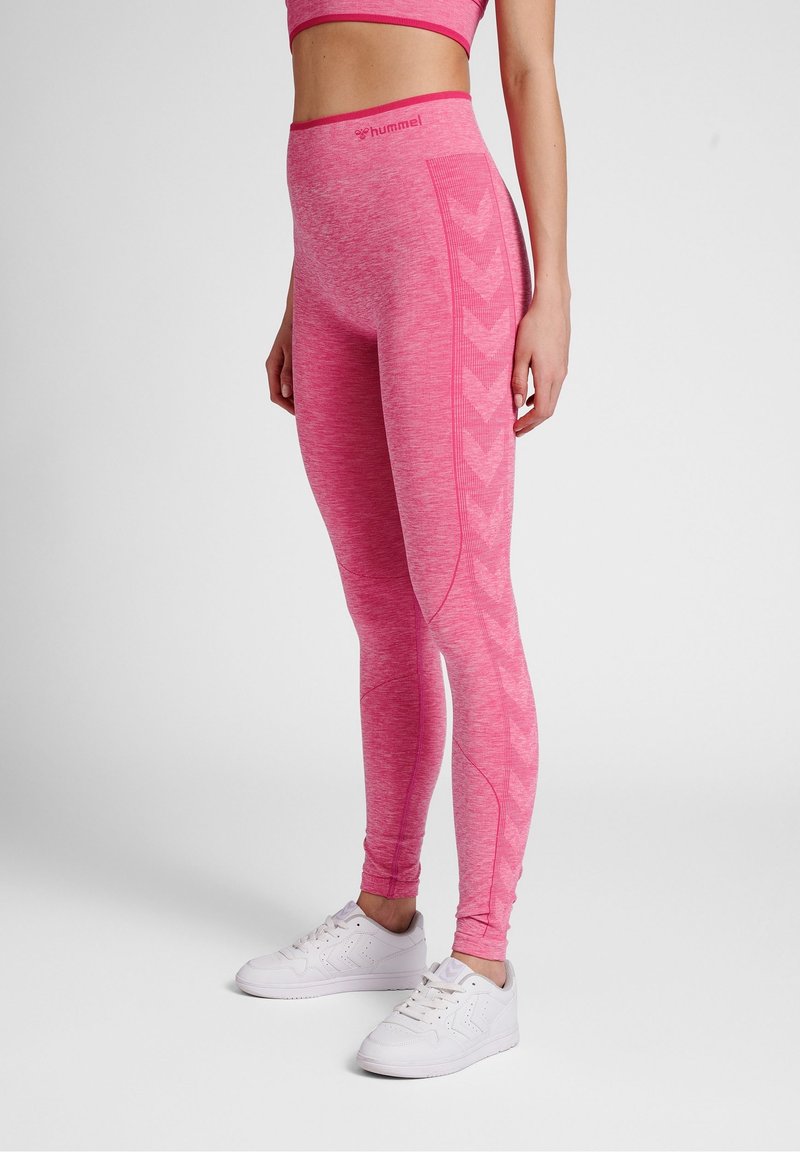 Růžové sportovní legíny s melírovanou texturou, zdobené postranními vzory ve tvaru šipek a vysokým pasem. Kombinovány s bílými teniskami.