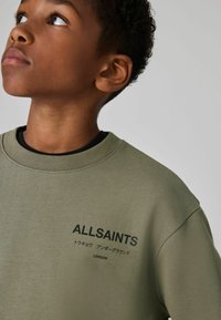 Maslinasto zeleni dukserica s rebrastim okovratnikom i crnim natpisom "ALLSAINTS" i "LONDON." Mekani materijal i udoban kroj.