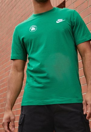 Mężczyzna w zielonej koszulce Nike z logo Celtics, stojący przy czerwonej ceglanej ścianie.