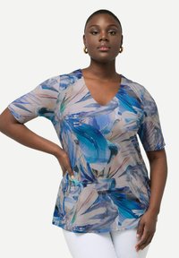 Top de manga corta con escote en V en tela transparente con un patrón floral azul, que presenta texturas en espiral y colores sutiles, combinado con pantalones blancos.