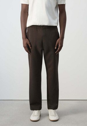 Persona con pantalones marrón oscuro, camisa blanca de manga corta y zapatillas blancas, de pie contra un fondo liso y claro.