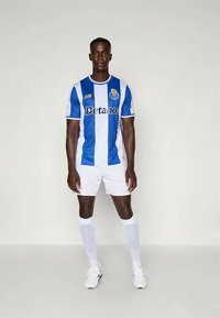 Kék és fehér csíkos focimez "Betano" logóval. Fehér rövidnadrággal, térd fölé érő fehér zoknival és sportcipővel párosítva.