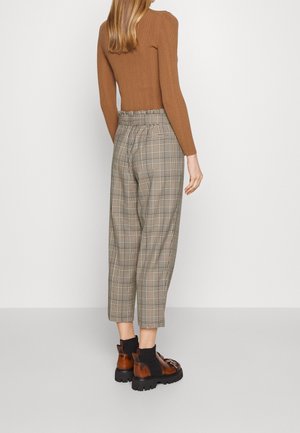 Femme portant un pantalon beige à carreaux taille haute, un haut marron côtelé à manches longues et des mocassins marron à semelle épaisse, debout devant un fond neutre.