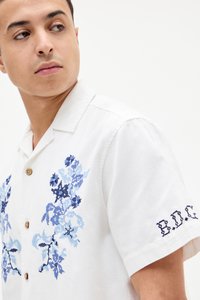 Cămașă albă cu mâneci scurte, decorată cu broderie florală albastră pe față și cu inscriptionarea "E.D.C." pe mânecă. Închidere cu nasturi din lemn.