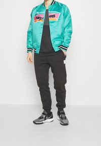 Chaqueta bomber de satén en color verde azulado con franjas negras y blancas, que presenta un logo gráfico colorido. Llevada sobre una camiseta oscura y pantalones de chándal grises.