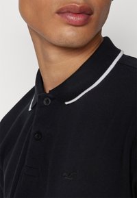 Polo noir en tissu texturé, avec un col blanc, trois boutons et un logo brodé sur la poitrine.