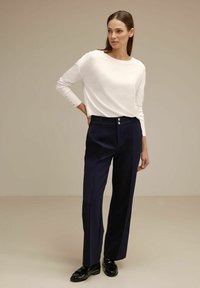 Top bianco a maniche lunghe con polsini a coste, abbinato a pantaloni blu navy a vita alta con due bottoni e una vestibilità rilassata. Scarpe nere completano l'outfit.