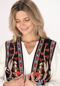 Schwarze bestickte Weste mit floralen Mustern in Rot, Blau und Grün. Getragen über einem weißen Oberteil mit V-Ausschnitt.