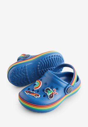 Zoccoli di gomma blu con suola decorata da un motivo arcobaleno, ornati con colorati charm decorativi rappresentanti un arcobaleno, un'auto e una bicicletta.