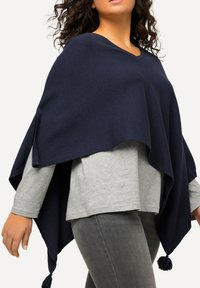 Laikkuva navy-punottu poncho V-kauluksella, jossa on kulmissa tupsut, on kerrostettu vaaleanharmaan pitkähihasen paidan ja harmaiden farkkujen päälle.