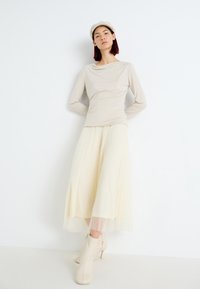 Beige langermet topp med samlet detalj, sammen med en plisserte, full-length lys gul skjørt og kremfargede ankelstøvler med tynn hæl.
