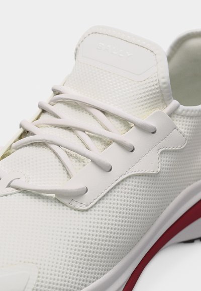 Baskets blanches texturées avec lacets blancs, une partie œillets en cuir, et un détail de semelle rouge et blanc, marquées "BALLY" sur la languette.