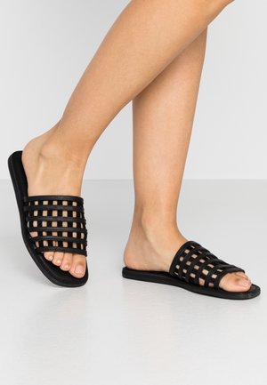 Stopy w czarnych klapkach na płaskiej podeszwie z wieloma poziomymi i pionowymi paseczkami, stojące na gładkiej jasnoszarej podłodze.