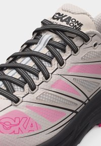 Prim-plan cu pantof de alergare gri deschis și roz, cu șireturi negre și logo-ul "HOKA ONE ONE" pe limba căptușită.