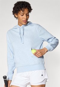 Felpa azzurra con una consistenza morbida, caratterizzata da una tasca frontale che contiene una pallina da tennis verde, indossata sopra pantaloni corti bianchi da atletica.