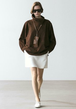 Frau trägt eine übergroße braune Jacke, weißen Minirock, weiße Schnürschuhe, schwarze Sonnenbrille und eine braune Tasche am Halsband.