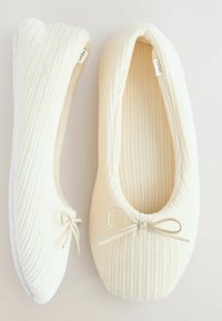 Ballerines de couleur crème en tissu à motifs texturés. Elles sont ornées d'un délicat nœud et présentent un design à bout rond. La semelle est blanche.