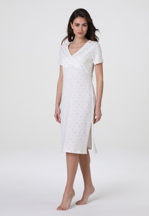 Donna in piedi a piedi nudi che indossa un vestito bianco a maniche corte, lungo fino al ginocchio, con piccoli motivi floreali e una spacco laterale.