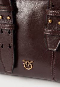 Σκούρο καφέ δερμάτινη τσάντα με διπλούς ιμάντες και μικρό χρυσό έμβλημα δύο πουλιών που σχηματίζουν κύκλο.