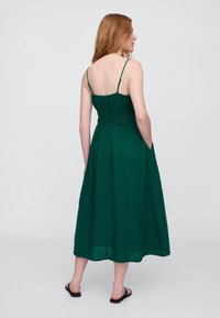 Zelené šaty po kolená s tenkými ramienkami, nazbieraným živôtikom a bočnými vreckami. Vyrobené z ľahkej tkaniny, so voľným strihom a štruktúrou.