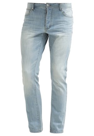 Mand i vinrød langærmet sweater, lyseblå opsmøgede jeans og hvide slip-on sko, der holder sort solbriller mod en ensfarvet lys baggrund.