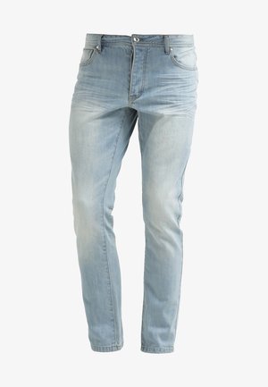 Mann in kastanienbraunem Langarmpullover, hellblauen hochgekrempelten Jeans und weißen Slip-On-Schuhen hält schwarze Sonnenbrille vor einfachem hellem Hintergrund.