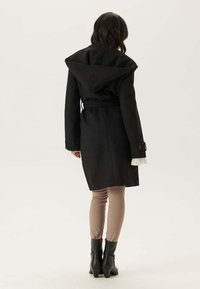 Donna che indossa un cappotto nero alla lunghezza del ginocchio con cappuccio, pantaloni beige e stivaletti neri, in piedi con la schiena rivolta verso l'osservatore.