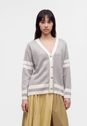 Jonge vrouw die een grijze gebuttoncardigan met witte strepen en een beige geplooide rok draagt, staand tegen een effen lichte achtergrond.