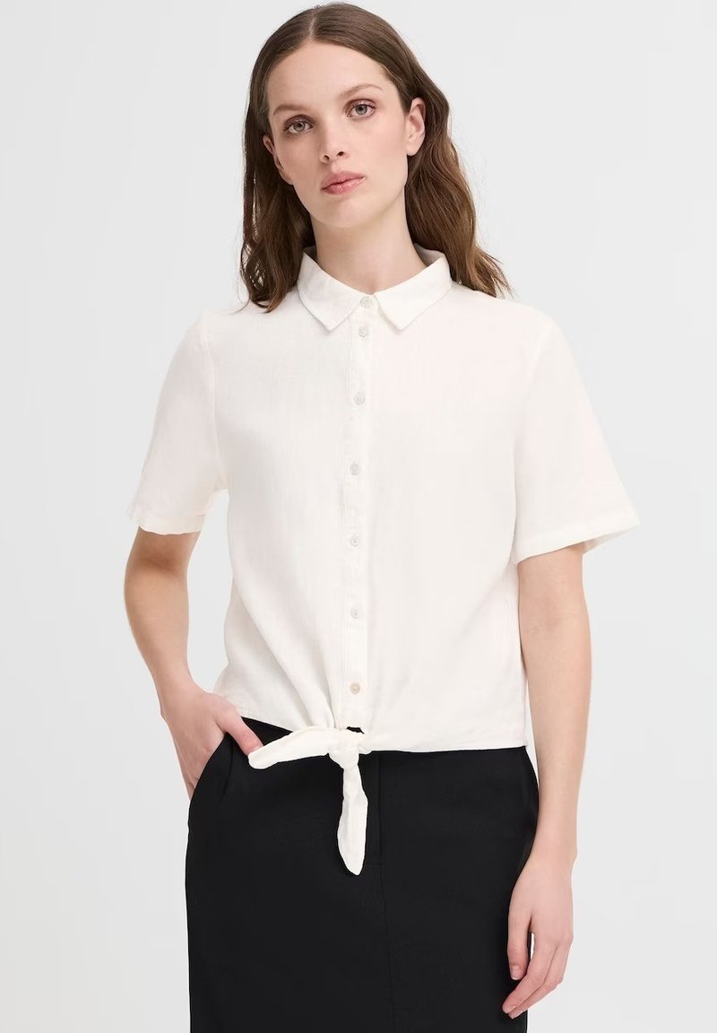 Camicia bianca a maniche corte con un design a bottoni e orlo anteriore annodato. Il tessuto sembra leggero e strutturato.