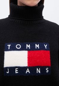 Pull à col roulé noir présentant un patch logo coloré avec des accents rouges, blancs et bleus, et un tissu tricoté texturé.