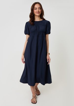 Woman wearing a navy blue midi dress with puff sleeves and pleated details, paired with brown strappy sandals and gold jewelry.