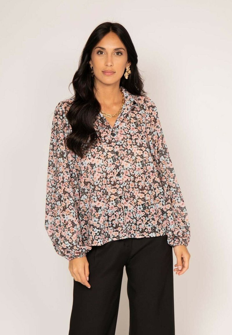 Blouse florale avec un fond noir et des fleurs roses, bleues et blanches. Manches bouffantes longues et fermeture à boutons devant.