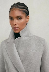 Sivi volneni plašč z visokim ovratnikom in teksturiranim tkanjem, z obliko reverja v obliki šala. Nošen preko črnega puli ovratnika.