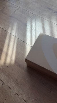 Caixa retangular bege sobre um chão de madeira com textura de grão visível. Sombras suaves projetadas pela luz do sol criam um padrão na superfície.