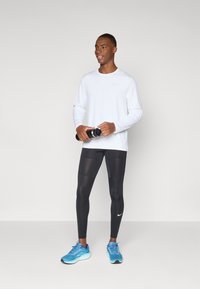 White long-sleeve shirt, black leggings, and blue athletic shoes. The shirt features a subtle logo; leggings have a fitted design and smooth texture.