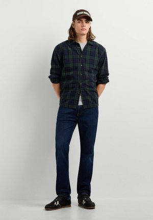 Jonge man met een bruin petje, donkergroen-blauw geruit overhemd, donkerblauwe spijkerbroek en zwarte sneakers, staand met de handen achter de rug.