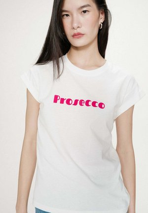 Jeune femme aux longs cheveux noirs portant un t-shirt blanc avec un texte rose vif "Prosecco" à l'avant, fond clair uni.