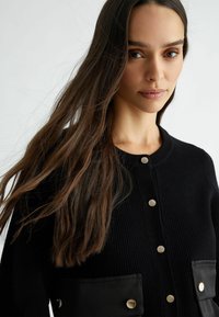 Cardigan nero a coste con bottoni a pressione argentati e due tasche frontali. Realizzato in tessuto testurizzato con una finitura morbida e scollatura pulita.