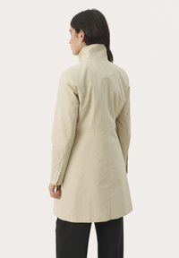 Beige trenchcoat med hög krage, långa ärmar och en figursydd form. Slätt tyg och rena linjer; ingen synlig hårdvara eller mönster.