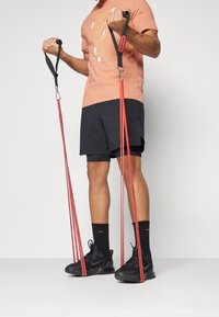 Man in zwarte shorts en sneakers traint met rode weerstandsbanden, terwijl hij de handgrepen omhoog trekt terwijl hij op de banden staat met zijn voeten.