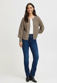 Cardigan marrón de punto con escote redondo, frente abierto y mangas largas ligeramente abullonadas. Lleva puesto sobre una camiseta blanca y jeans azules.