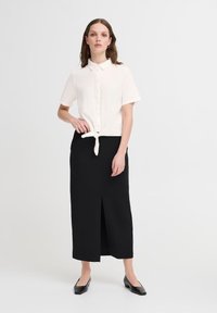 Camicia bianca a maniche corte con bottoni legata in vita, abbinata a una gonna nera al ginocchio con una fessura frontale e scarpe flat nere.
