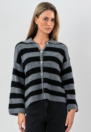 Žena nosi sivý a čierny pletený kardigán so širokými horizontálnymi pruhmi, zapínaný na gombíky, s dlhými rukávmi a golierom.