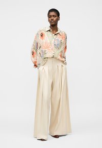 Žena v béžových kalhotách s širokými nohavicemi a volné košili s květinovým potiskem, stojící před jednobarevným světlým pozadím.