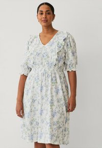 Robe blanche avec un motif floral bleu et vert, manches courtes à volants, décolleté en V, bordure froncée et taille smockée. Texture de tissu léger.