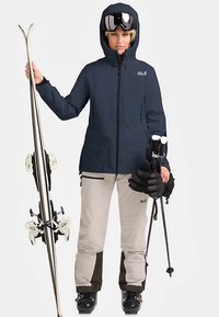 Veste imperméable marine avec capuche, pantalon de ski beige et noir, gants noirs, bâtons de ski et skis, présentant un design élégant et des accents fonctionnels.