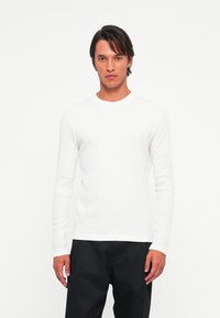 Man in een eenvoudige witte lange mouw shirt en zwarte broek, staand tegen een witte achtergrond, recht vooruit kijkend met een neutrale uitdrukking.
