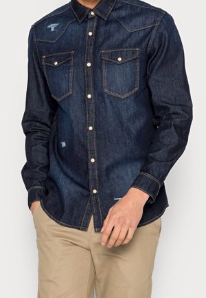 Homme portant une chemise en jean bleu foncé boutonnée avec des poches poitrine et un pantalon beige, mains partiellement dans les poches.